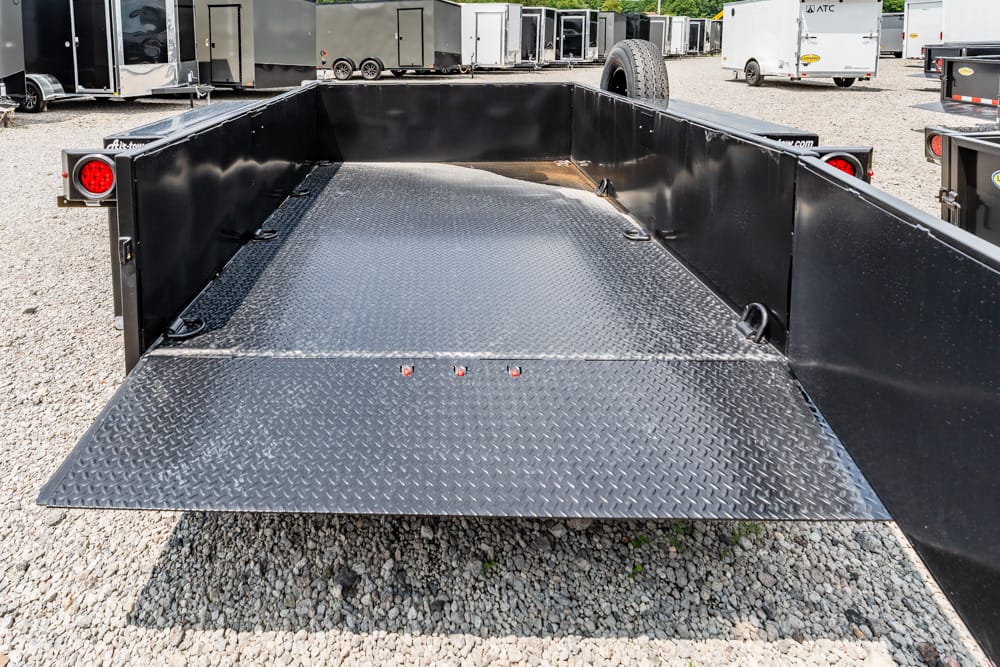 Black flatbed trailer with a diamond-plate ramp lowered to the ground in a gravel lot, with other trailers in the background.