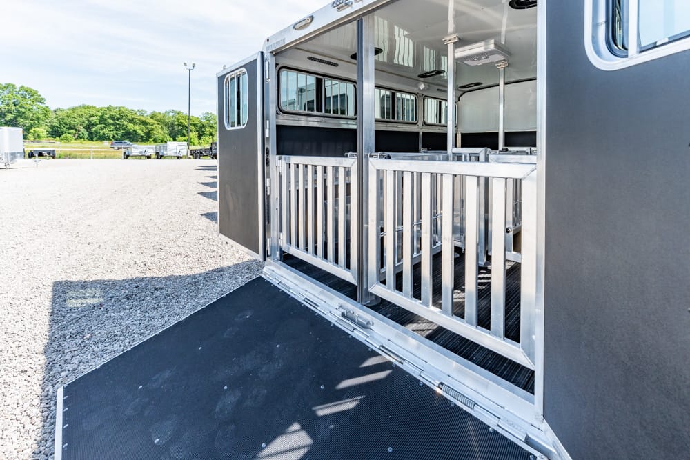 Open horse trailer with metal dividing stalls, ramp down, on a gravel lot with other trailers in the background.
