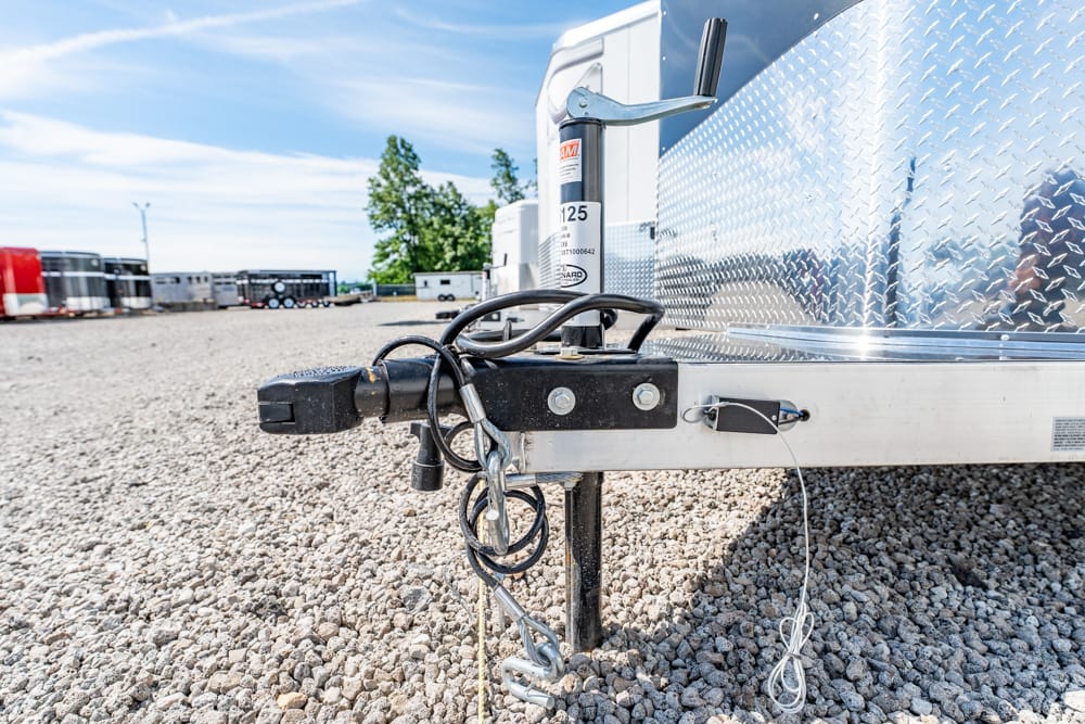 Close-up of a trailer hitch and jack on a gravel lot with a chrome diamond-plate trailer side in the background.