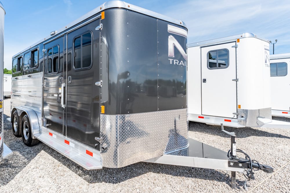 Black two-axle horse trailer with diamond-plate front hitching area, parked on gravel with a white trailer nearby.
