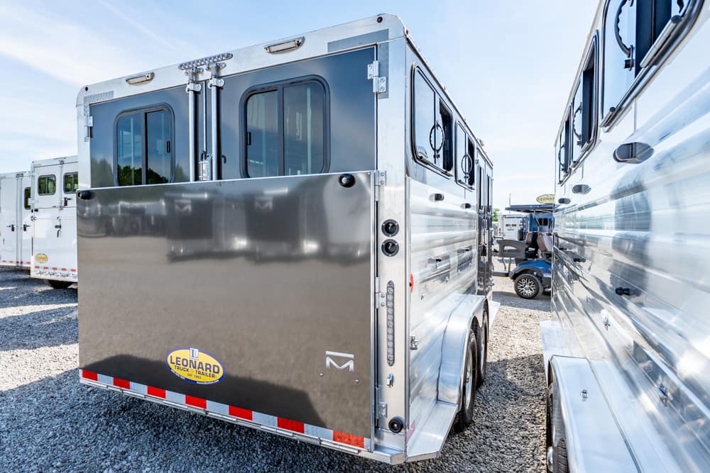 Shiny silver horse trailer parked between other trailers in a gravel lot, Leonard brand logo visible on the rear door
