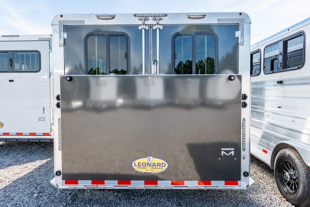 Back of a shiny dual-door horse trailer with a Leonard trailer logo on the lower panel, parked on gravel with neighboring trailers nearby.