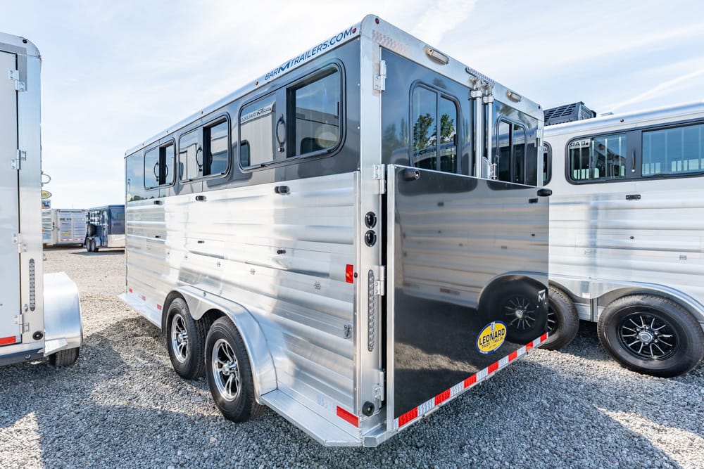 Silver aluminum horse trailer with rear doors open in a gravel lot, other trailers parked nearby.