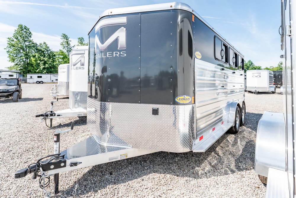 Black and silver two-axle travel trailer with diamond-plate nose, hitch in foreground, parked on a gravel lot.