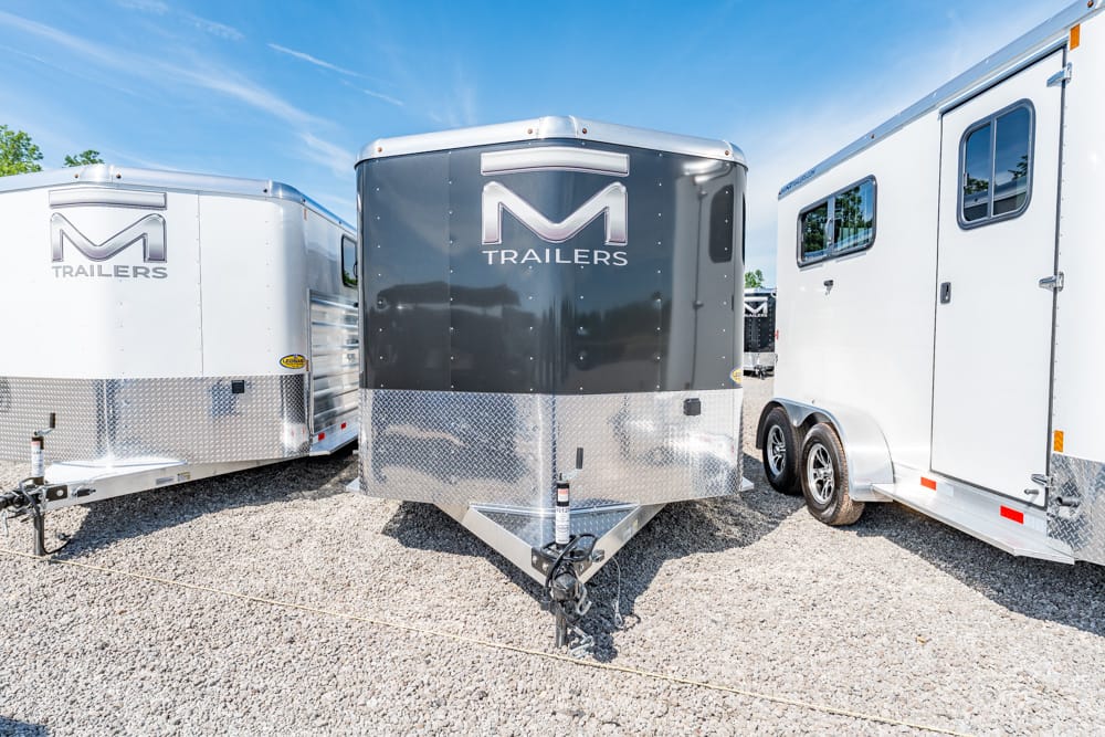 Front view of a dark gray M Trailers trailer between two white trailers on a gravel lot under a blue sky.