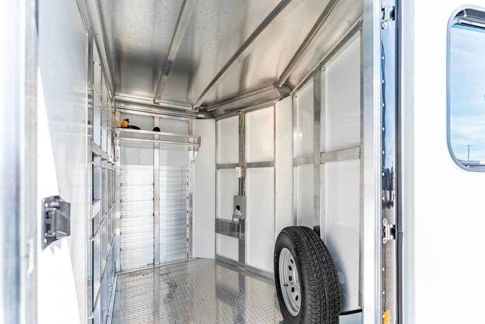 Empty aluminum trailer interior with metal shelves on the left, a closed rear door, and a mounted spare tire on the right wall.