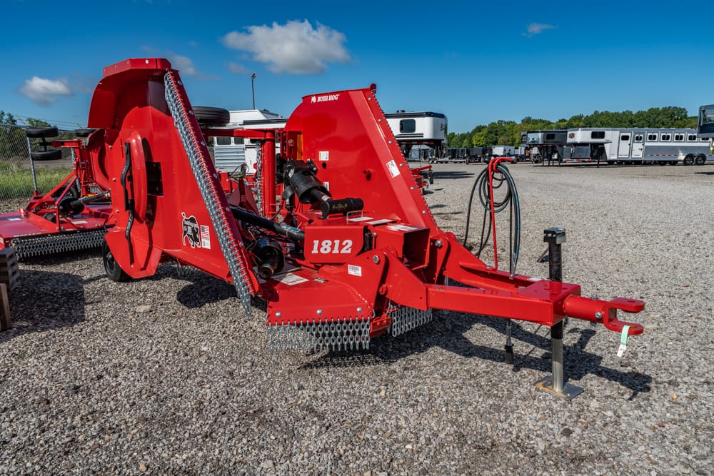Red Bush Hog 1812 rotary cutter attached to a hitch, parked on a gravel lot with other farming equipment visible in the background.