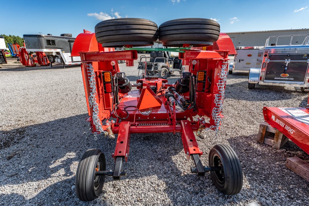 Rear view of a red agricultural implement with dual top wheels and chain guards, parked on a gravel lot behind trailers