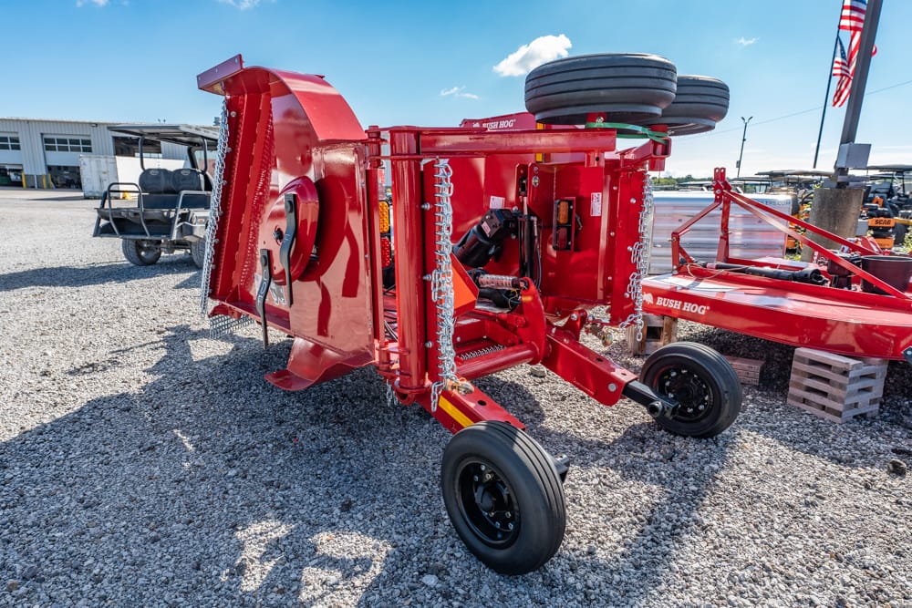 Red Bush Hog rotary mower attachment on a gravel lot, with wheels, chains, and a hitch ready for transport.