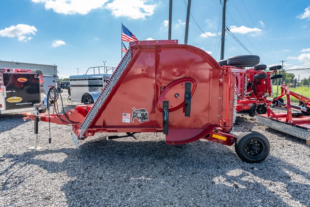 Red round-baler (Bush Hog) parked on a gravel lot, with other equipment and an American flag in the background.