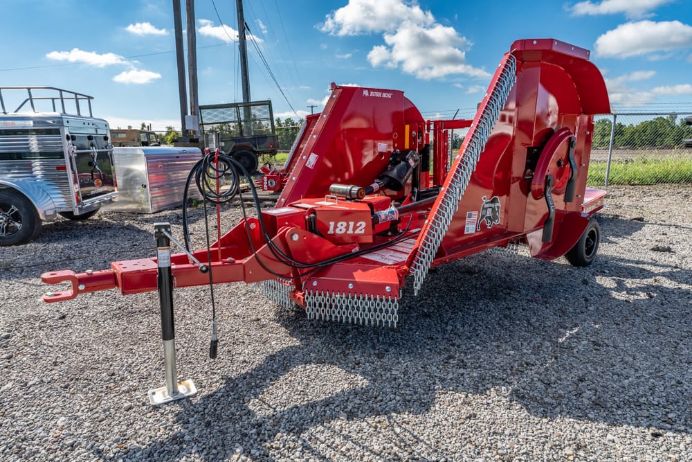 Red Bush Hog rotary cutter/trailer attachment with hydraulic hoses and chain guards, parked on a gravel lot.