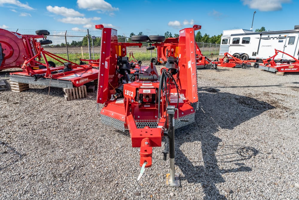 Front view of red agricultural machinery on a trailer in a gravel lot, with a chain-link fence and a white camper in the background.