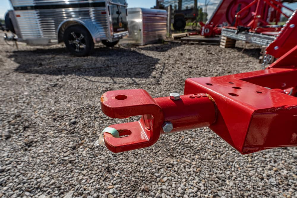 Red trailer hitch coupler with a locking pin on a gravel lot, prep for connection to a tow vehicle; equipment in the background.
