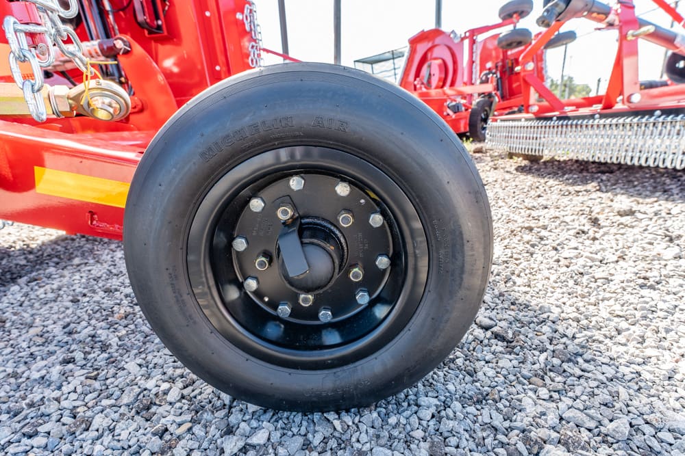 Close-up of a Michelin tire on a black wheel with a bolt hub, attached to red agricultural equipment on gravel, with other red gear in the background.