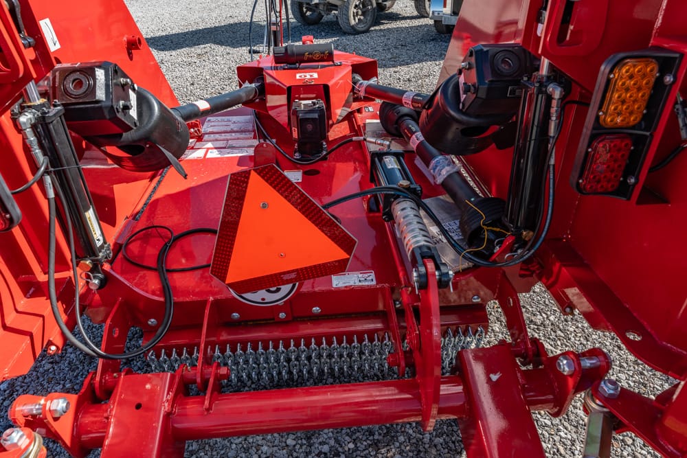 Close-up of red agricultural equipment showing hydraulic hoses, metal linkages, and a reflective orange triangle warning sign on a gravel surface