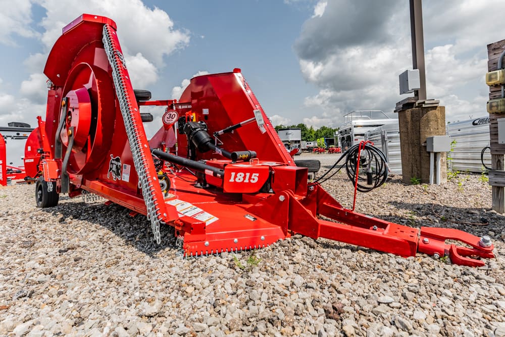 Red agricultural baler on a gravel lot, with hoses and gear exposed and other equipment in the background (display at a sales yard).