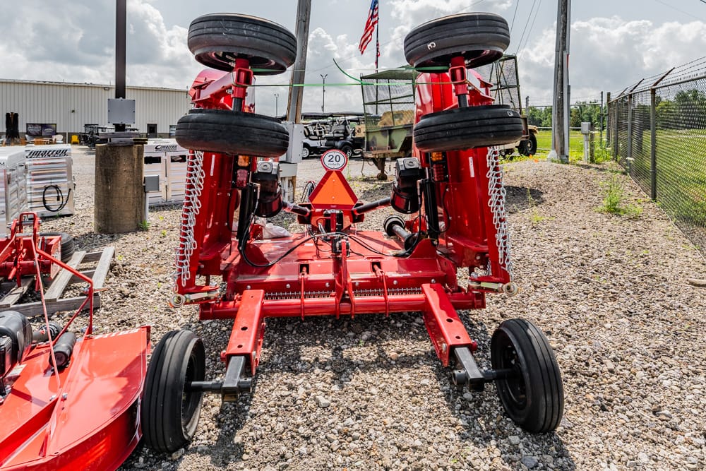 Red agricultural implement lying on a gravel lot, wheels upside down on the frame, with other equipment and a fence in the background.