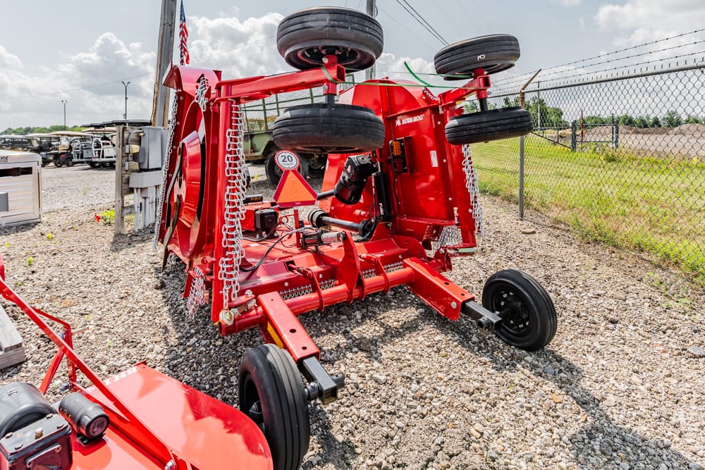Red agricultural implement lying on its side in a gravel lot, with wheels and chains visible and a caution triangle sign attached nearby near a chain-link fence.