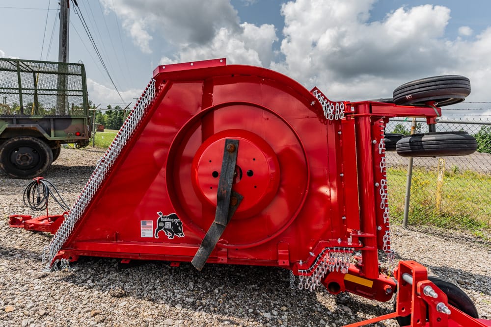 Red Bush Hog rotary cutter/brush mower on a transport trailer, visible chain guards and drivetrain on gravel.