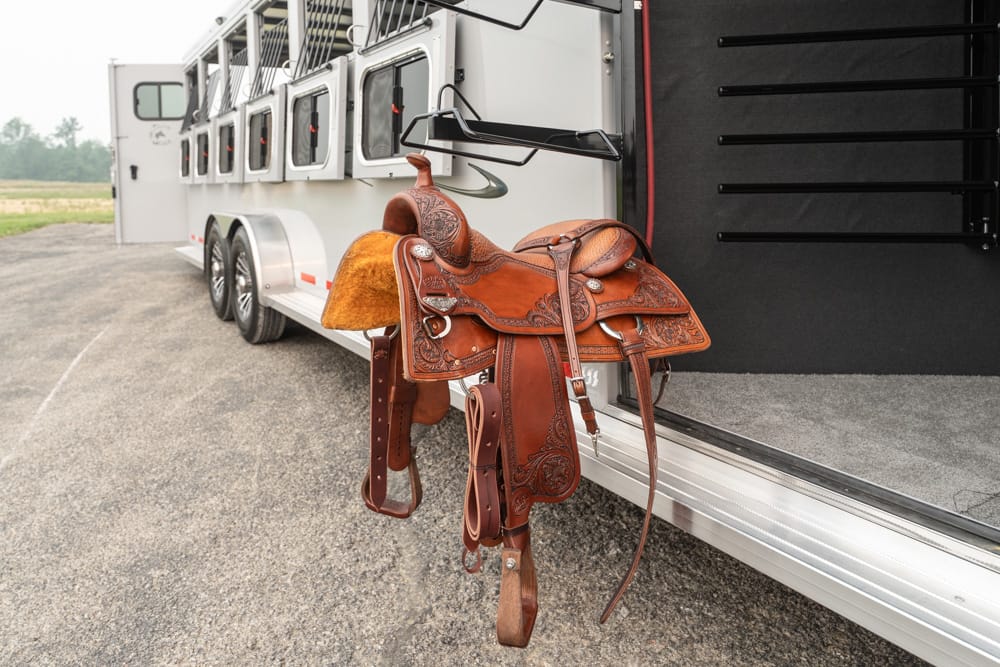 A brown leather Western saddle hangs on the open side of a white horse trailer, ready for use in the trailer stalls.