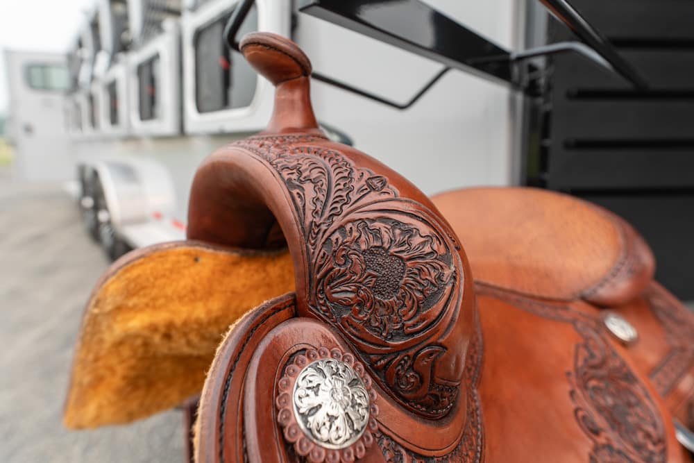 Close-up of a brown leather Western saddle with intricate floral tooling and a silver concho, against a blurred trailer in the background.
