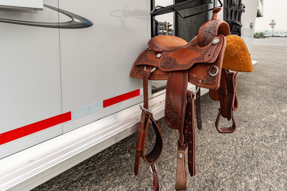 Western leather saddle with ornate tooling hanging on the side of a light-colored trailer.