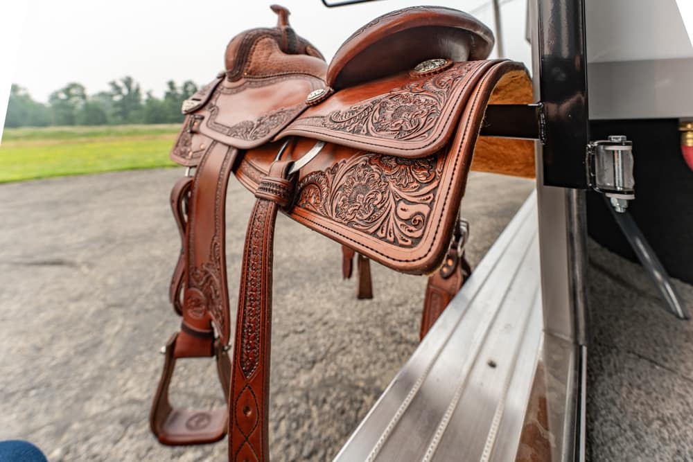 Decorative brown Western saddle with floral tooling resting on a trailer step beside an asphalt surface.