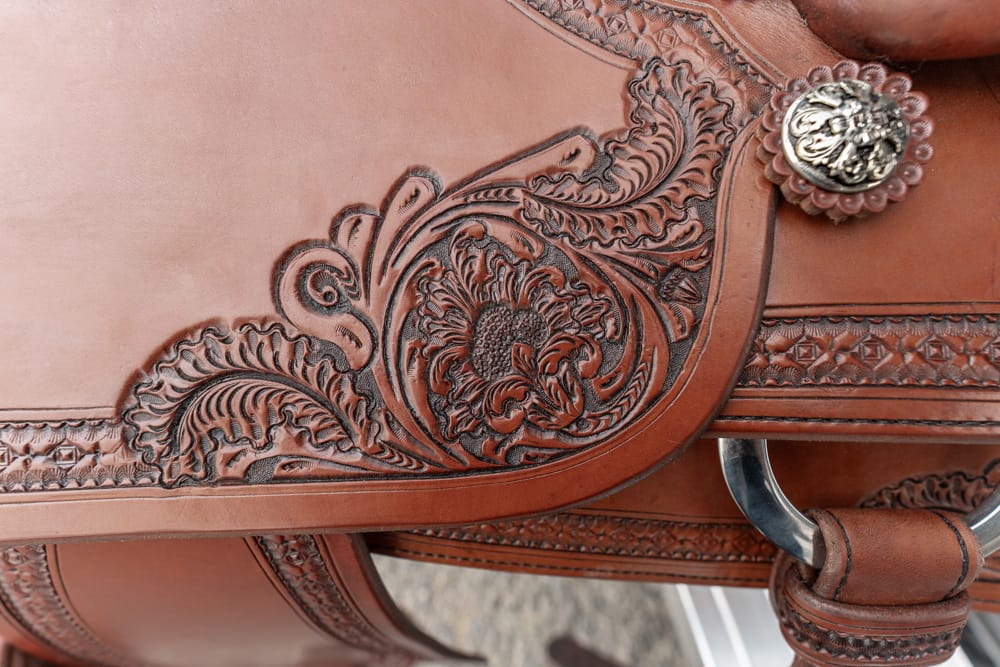 Close-up of a brown leather saddle with ornate floral tooling and a silver concho in the upper right corner.