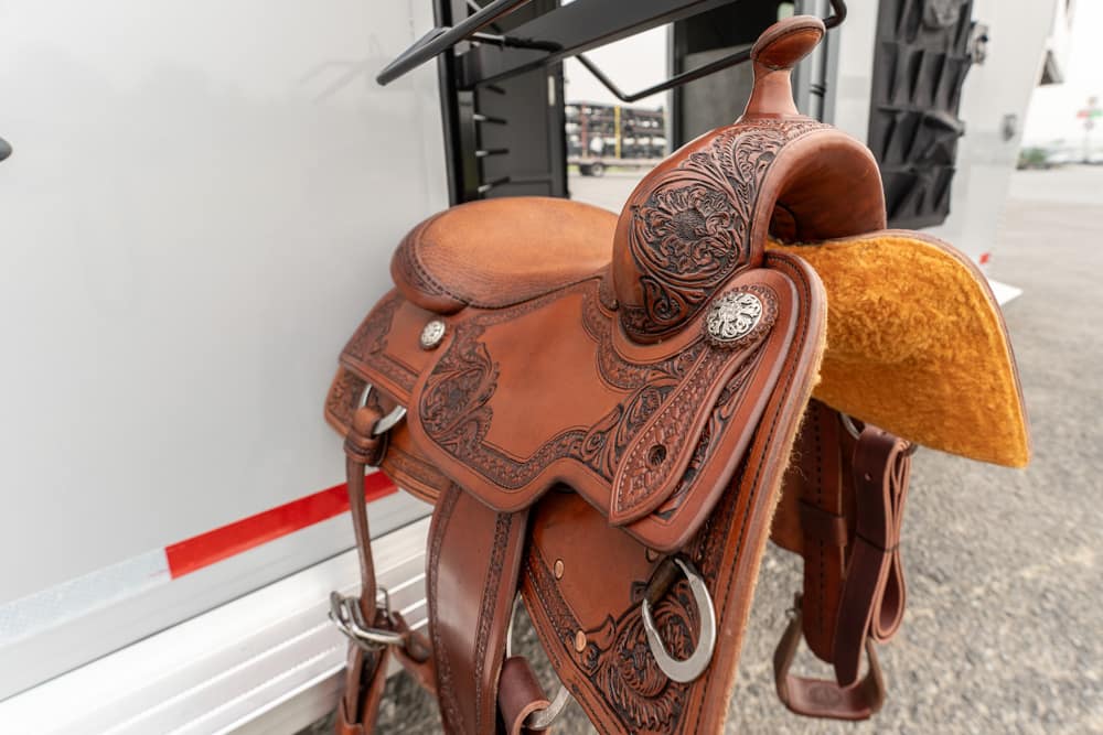 Leather western saddle with floral tooling resting on a trailer hitch, showing detailed craftsmanship and a suede seat pad nearby