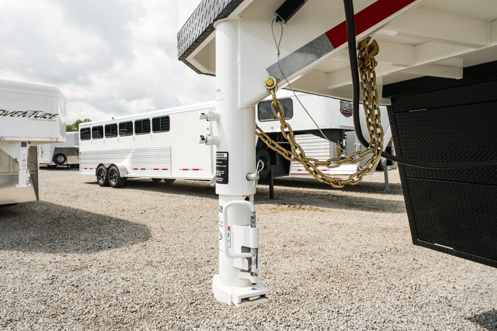 Close-up of a fifth-wheel trailer’s hitch and safety chains on a gravel lot, with other trailers in the background.