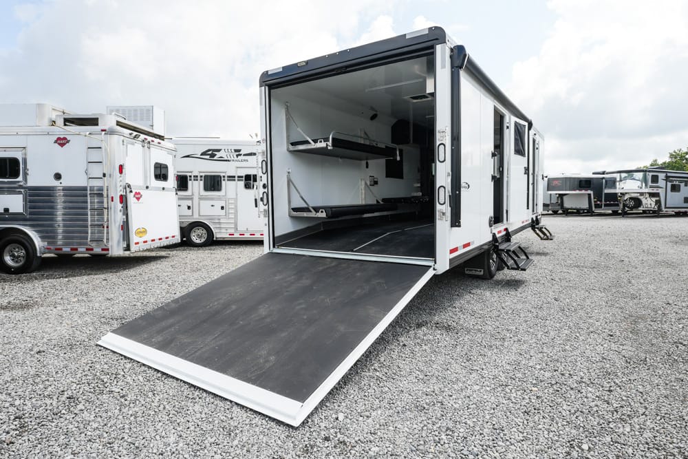 Large white toy hauler trailer with ramp down, exposing interior storage shelves, parked on a gravel lot with other trailers.