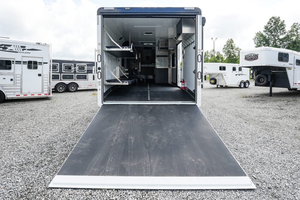 Open rear of a large cargo trailer with the ramp down, showing interior shelves and equipment on a gravel lot with other trailers nearby.