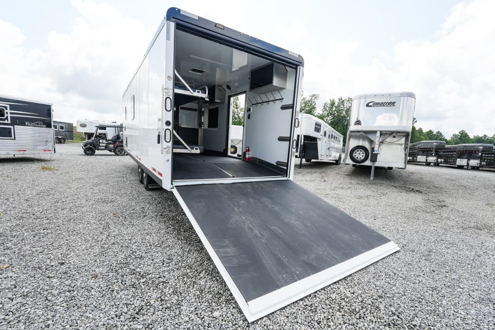 White cargo trailer with its ramp lowered for loading, parked on a gravel lot among other trailers; interior compartments visible through the open rear doors.