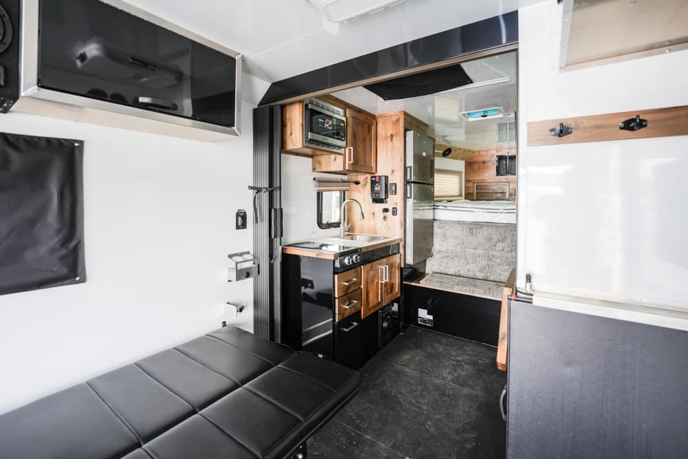 Interior of a modern camper van with a black sofa, wooden kitchen cabinets, sink, stove, and compact fridge next to a sleeping area.