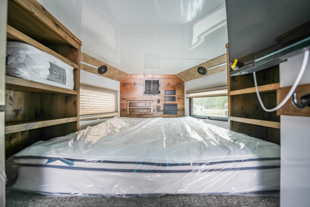 Interior of a camper bedroom with a large bed, wood shelves on both sides, and a back wall feature panel with vents.