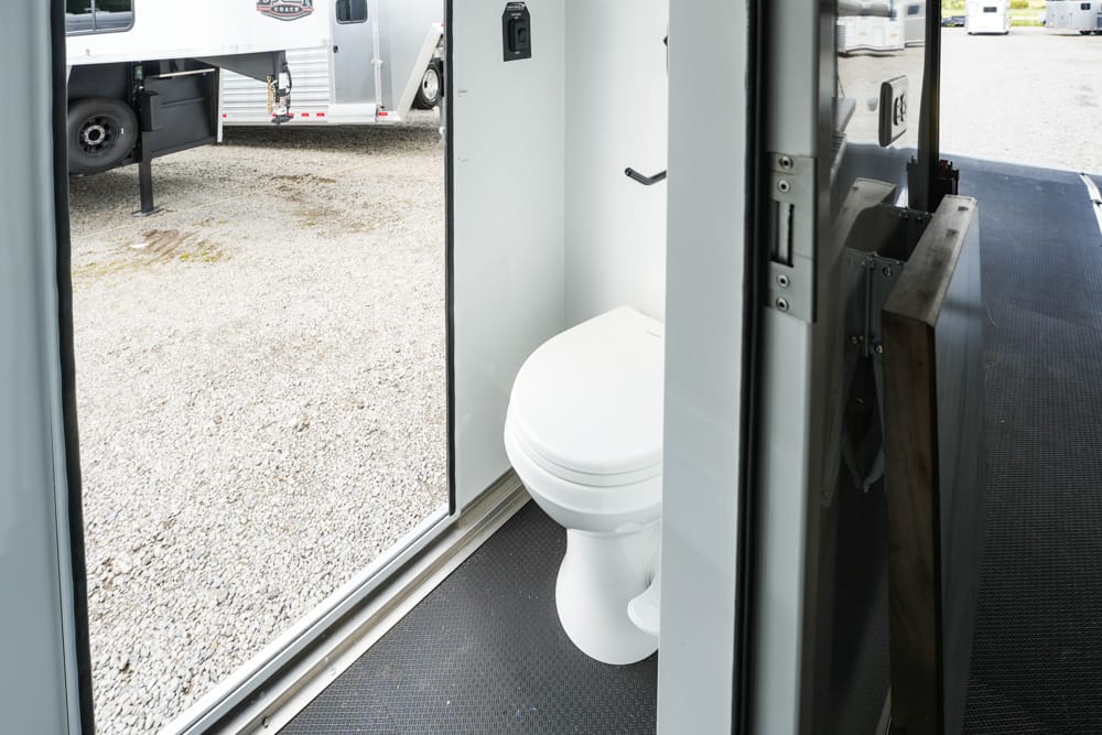 Interior of a portable toilet trailer with a white toilet visible through an open doorway, gravel ground outside and other trailers in the lot