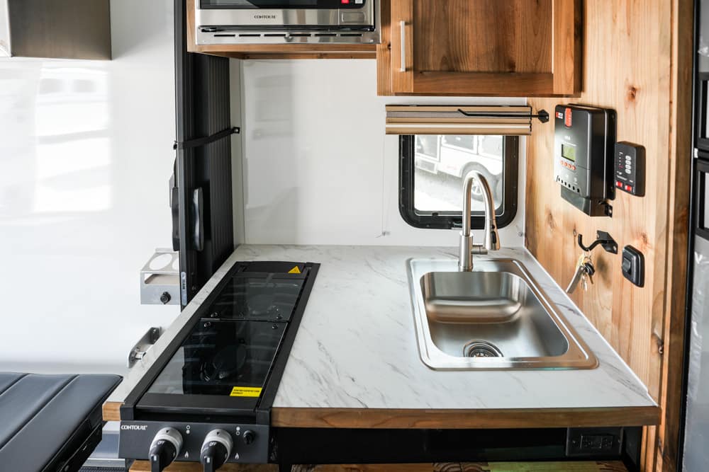 Compact camper kitchen with a stainless steel sink, two-burner stove, marble-pattern counter, and wooden cabinets above.