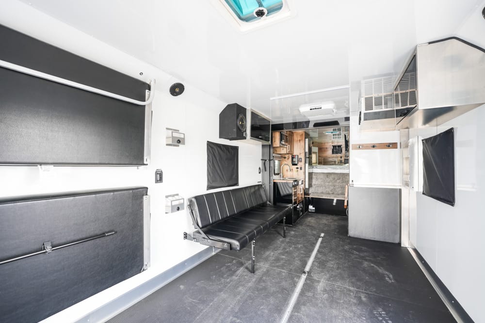 Interior of an ambulance with black bench seating along the left wall, white walls, and cabinets toward the rear, conveying a patient-care space.