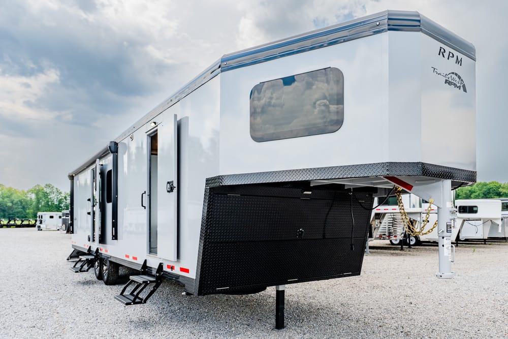 White fifth-wheel travel trailer parked on a gravel lot, with exterior stairs and a large side window visible under a cloudy sky.