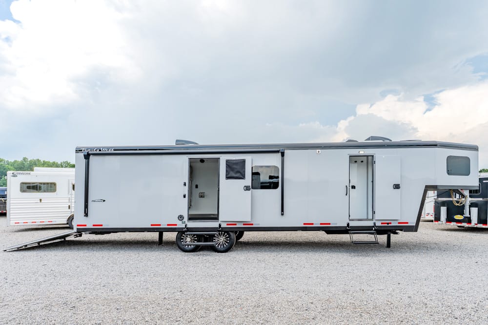 Long silver travel trailer parked on a gravel lot, with exterior doors open and a ramp at the left end for loading or accessibility features