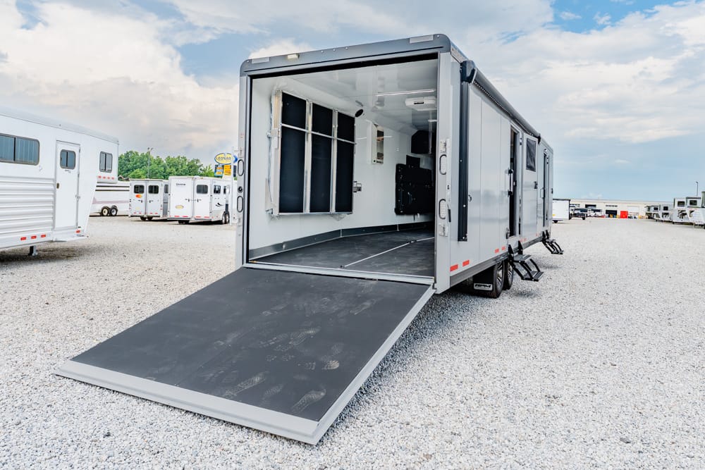 Silver trailer with the rear ramp down, revealing an empty interior on a gravel lot at a vehicle yard.