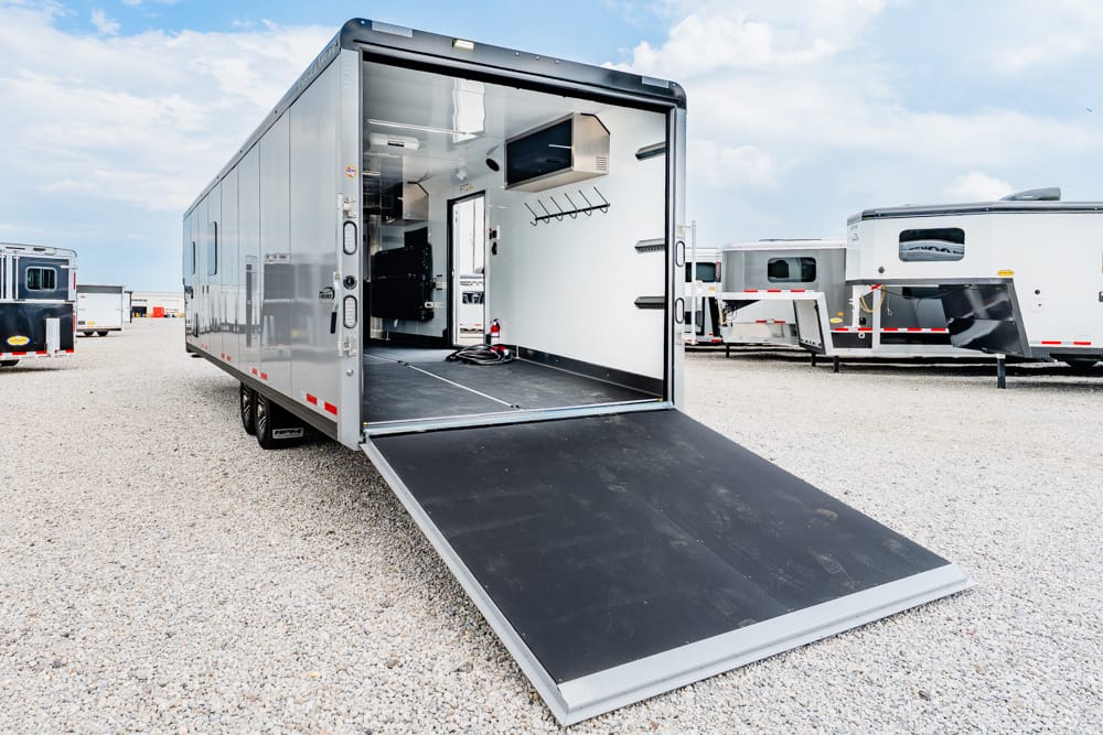 Silver cargo trailer with rear ramp lowered, exposing an empty interior ready for loading on a gravel lot.