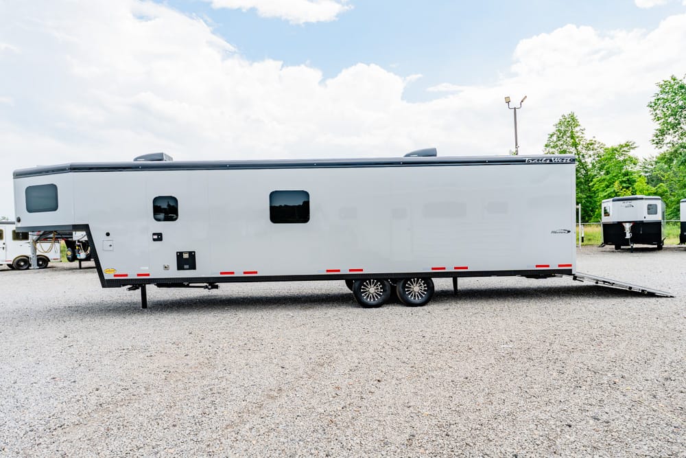Large gray travel trailer with two axles on a gravel lot; rear ramp extended when parked.