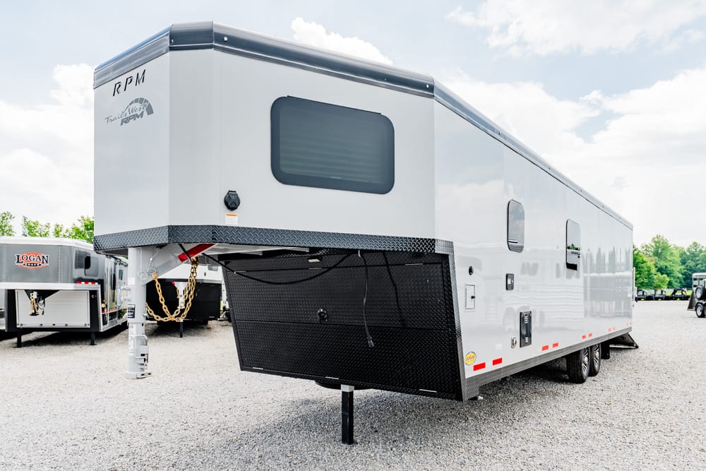 White fifth-wheel trailer with extended hitch and black diamond-plate storage box at the front, parked on gravel in a dealership lot.