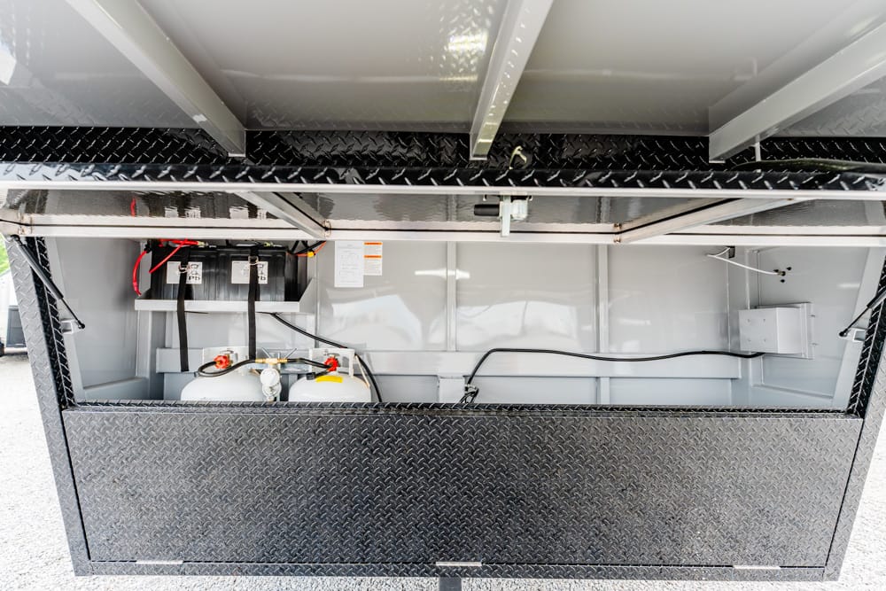 Interior of a diamond-plate utility trailer storage bay with two white propane tanks secured inside, connected by hoses and gauges, and electrical wiring visible.