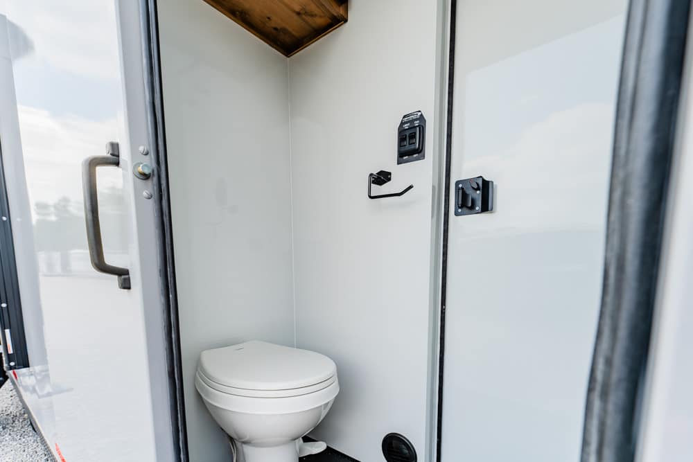 Interior of a small portable toilet trailer with a white toilet and pale walls, door open.
