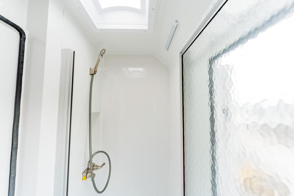 White shower stall with a handheld showerhead and hose mounted on the wall, frosted glass window to the right, and a skylight above.