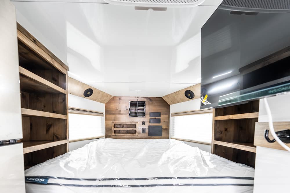 Interior of a camper bedroom with a centered bed, white cover, wooden shelves on both sides and built-in storage above.