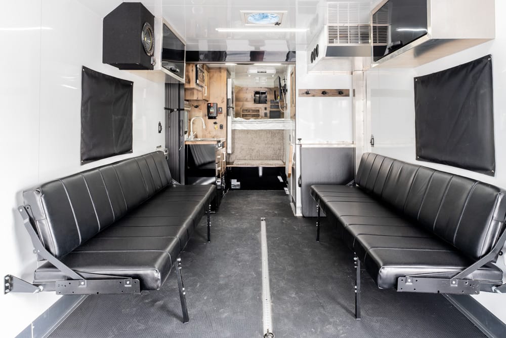 Interior of an ambulance with two long black bench seats along the sides, white walls, and medical equipment toward the rear.