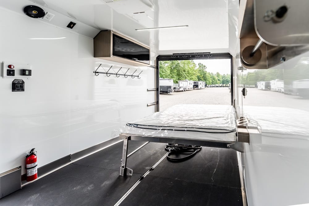Interior of a mobile medical trailer with a metal gurney covered in plastic, wall hooks, shelves, and a fire extinguisher; open rear door shows parked trailers outside.